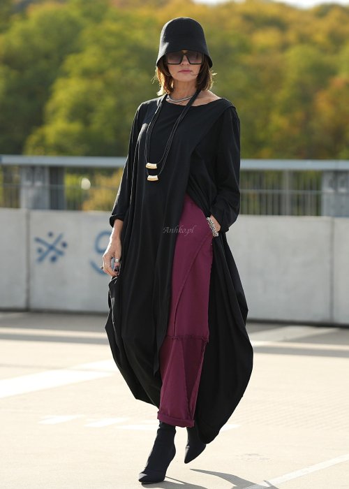Czarna sukienka maxi o asymetrycznym dole i luźnym kroju oversize. Modelka prezentuje stylizację z czarnymi botkami, kapeluszem i minimalistyczną biżuterią w miejskim stylu.