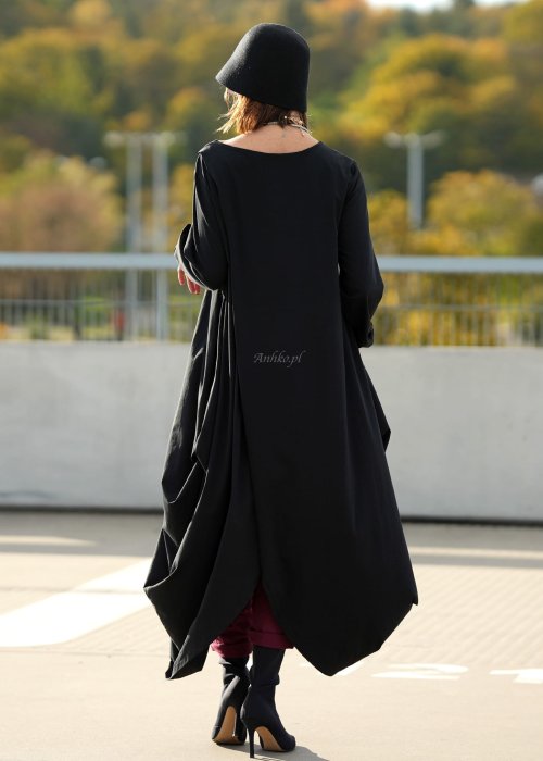 Czarna sukienka maxi o asymetrycznym dole i luźnym kroju oversize. Modelka prezentuje stylizację z czarnymi botkami, kapeluszem i minimalistyczną biżuterią w miejskim stylu.