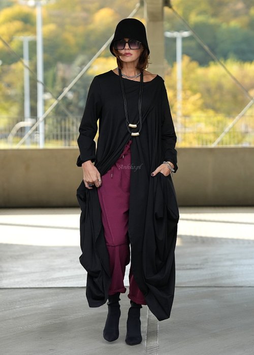 Czarna sukienka maxi o asymetrycznym dole i luźnym kroju oversize. Modelka prezentuje stylizację z czarnymi botkami, kapeluszem i minimalistyczną biżuterią w miejskim stylu.