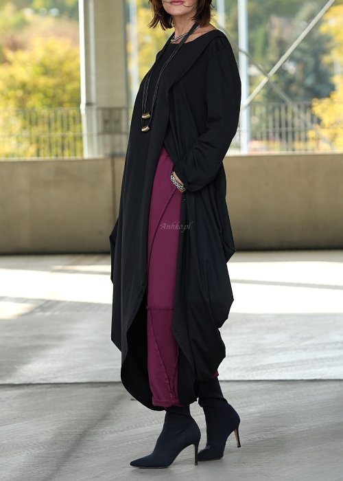 Czarna sukienka maxi o asymetrycznym dole i luźnym kroju oversize. Modelka prezentuje stylizację z czarnymi botkami, kapeluszem i minimalistyczną biżuterią w miejskim stylu.