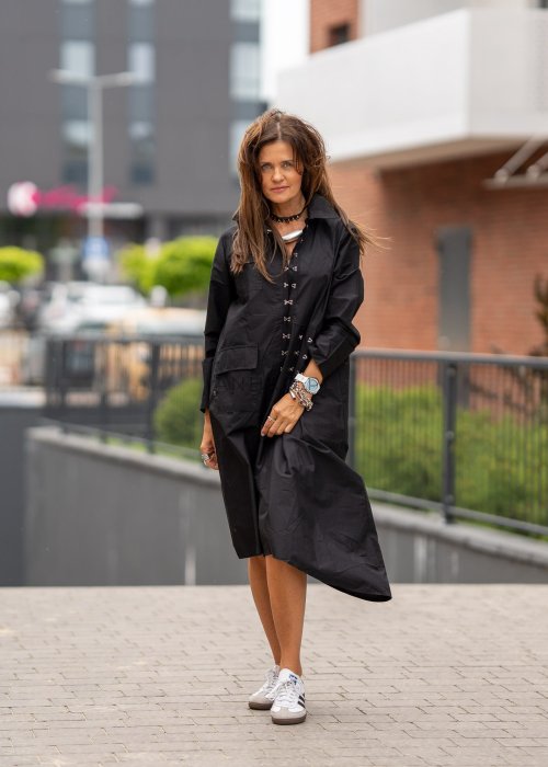 Czarna sukienka koszulowa midi z długim rękawem i ozdobnymi metalowymi zapięciami z przodu. Model pokazany w miejskiej stylizacji ze sneakersami, z wyraźnie zaznaczonym kołnierzykiem i luźnym fasonem.