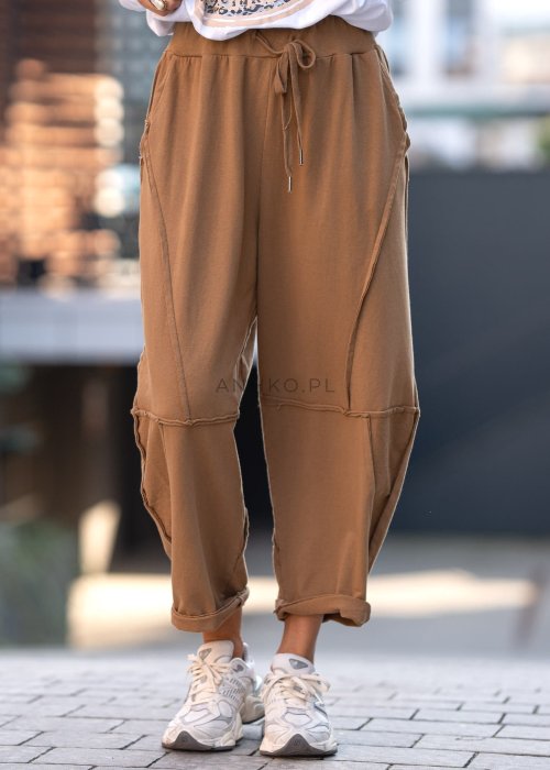 Damskie spodnie dresowe w kolorze camel z fantazyjnymi przeszyciami i elastycznym pasem, stylizowane z jasnymi sneakersami w miejskiej scenerii.