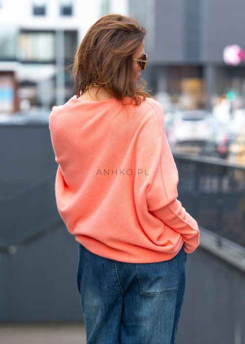 Łososiowy sweter damski oversize o kroju nietoperzowym z dekoltem łódka i asymetrycznym dołem, miejska stylizacja z jeansem.