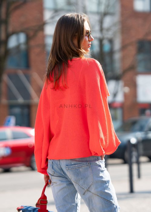 Koralowy sweter oversize z asymetrycznym dołem, stylizowany z jasnymi jeansami i czerwonymi dodatkami w miejskim looku.