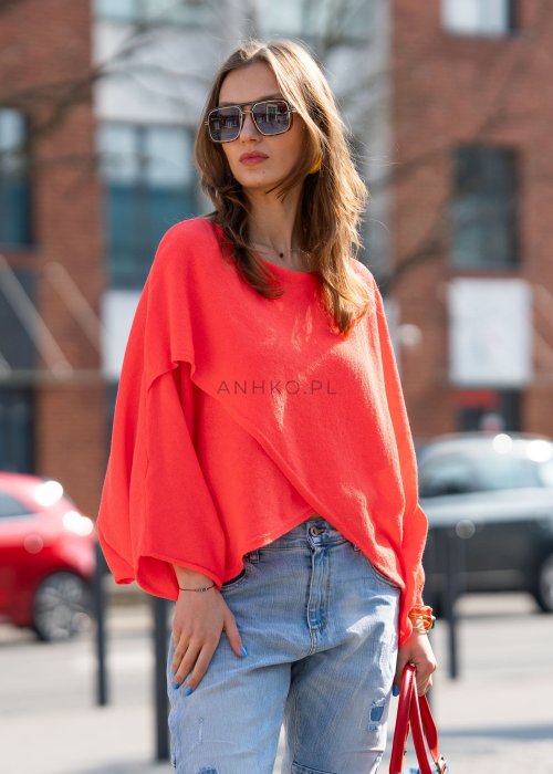 Koralowy sweter oversize z asymetrycznym dołem, stylizowany z jasnymi jeansami i czerwonymi dodatkami w miejskim looku.