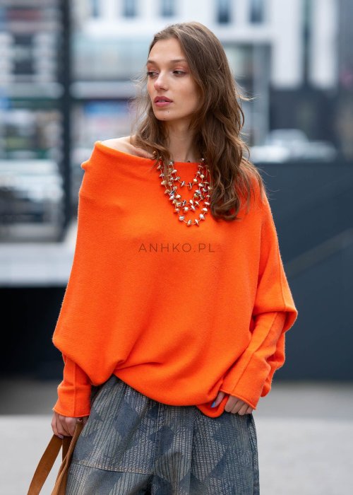 Pomarańczowy sweter damski oversize o nietoperzowym kroju z asymetrycznym dołem, stylizacja miejska z biżuterią i spodniami.