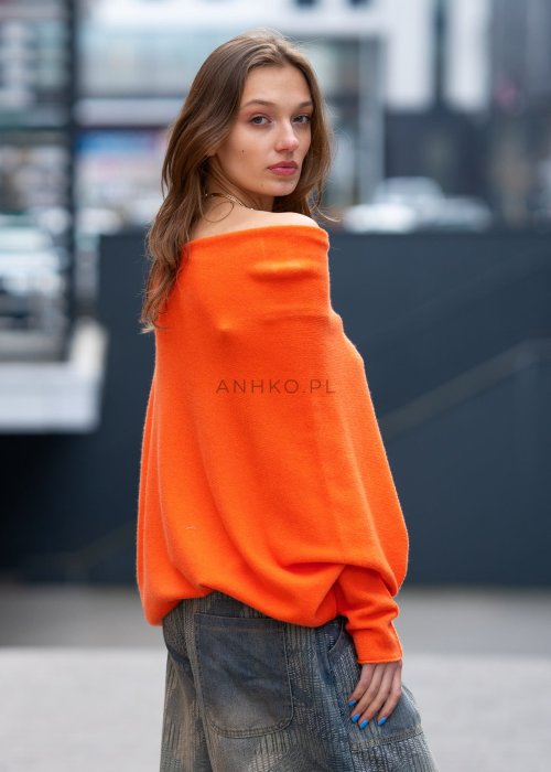 Pomarańczowy sweter damski oversize o nietoperzowym kroju z asymetrycznym dołem, stylizacja miejska z biżuterią i spodniami.