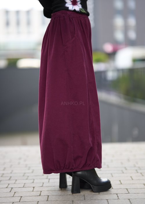 Śliwkowa spódnica maxi z elastyczną talią i kieszeniami, o luźnym kroju, zestawiona z czarną bluzą i czarnymi botkami na obcasie w miejskiej stylizacji.