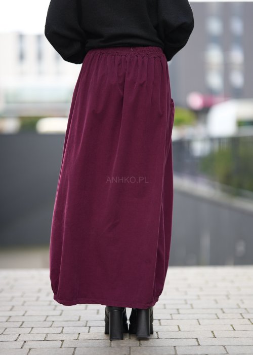 Śliwkowa spódnica maxi z elastyczną talią i kieszeniami, o luźnym kroju, zestawiona z czarną bluzą i czarnymi botkami na obcasie w miejskiej stylizacji.