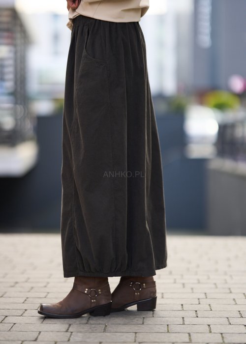 Brązowa spódnica maxi z elastyczną talią i kieszeniami, o luźnym kroju, zestawiona z beżową bluzką i brązowymi botkami w eleganckiej, miejskiej stylizacji.