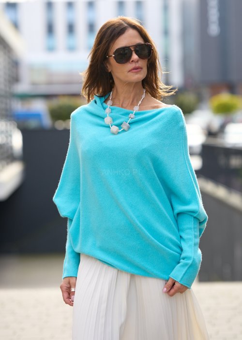 Jasnoniebieski sweter oversize o nietoperzowym kroju z dekoltem łódka i asymetrycznym dołem zestawiony z jasną plisowaną spódnicą oraz dużymi okularami przeciwsłonecznymi.