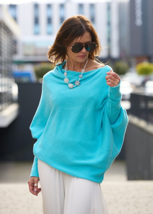 Jasnoniebieski sweter oversize o nietoperzowym kroju z dekoltem łódka i asymetrycznym dołem zestawiony z jasną plisowaną spódnicą oraz dużymi okularami przeciwsłonecznymi.
