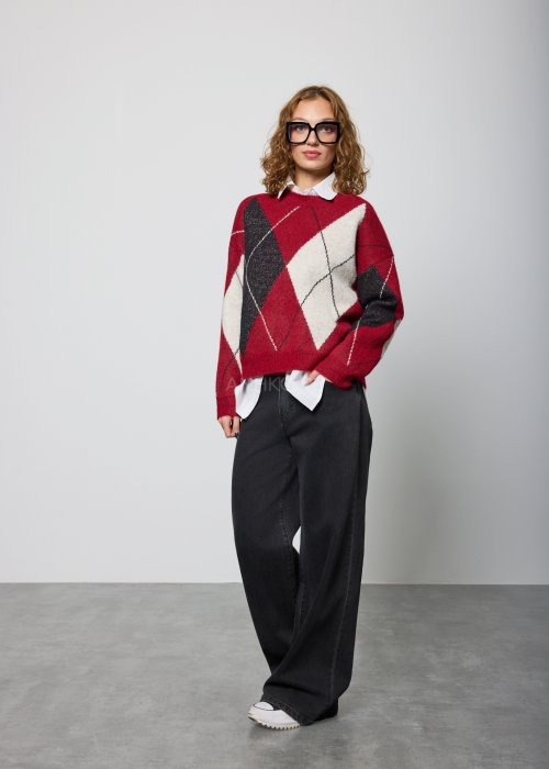 Damski sweter w romby w odcieniach czerwieni, czerni i ecru, o luźnym kroju i długich rękawach, noszony na białej koszuli z kołnierzykiem, zestawiony z czarnymi jeansami.