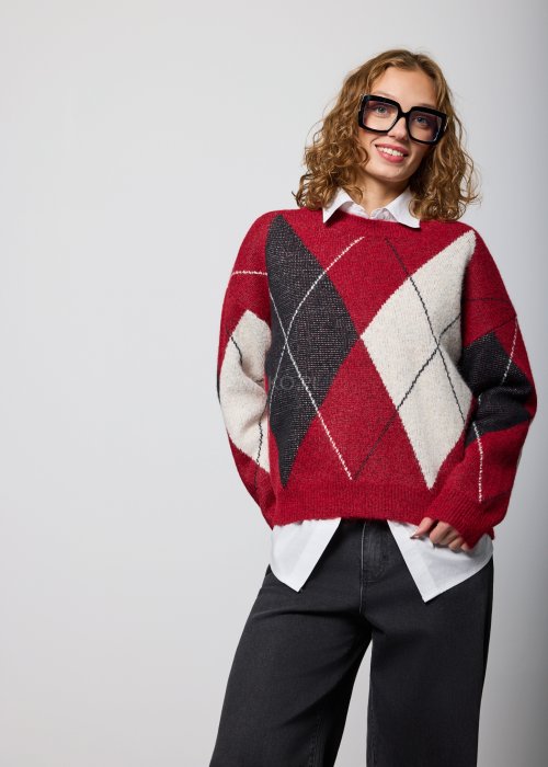 Damski sweter w romby w odcieniach czerwieni, czerni i ecru, o luźnym kroju i długich rękawach, noszony na białej koszuli z kołnierzykiem, zestawiony z czarnymi jeansami.