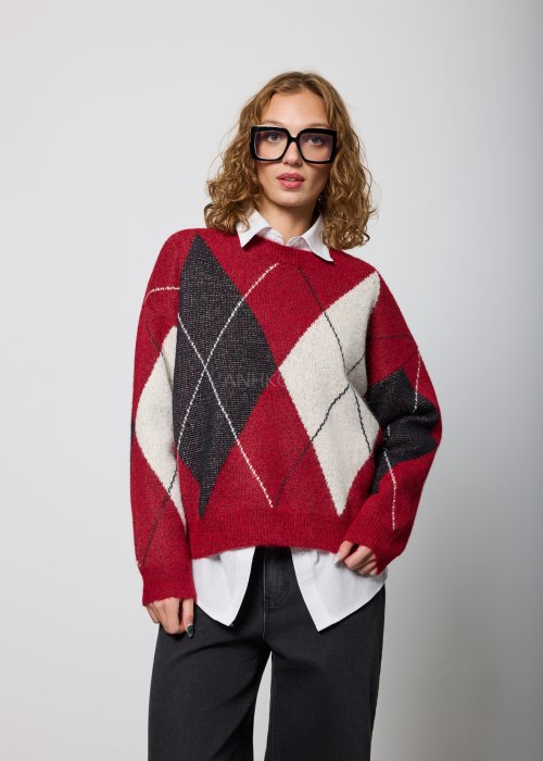 Damski sweter w romby w odcieniach czerwieni, czerni i ecru, o luźnym kroju i długich rękawach, noszony na białej koszuli z kołnierzykiem, zestawiony z czarnymi jeansami.