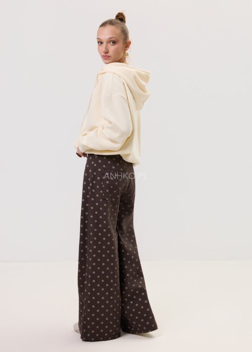 Brązowe damskie spodnie Retro Dots z szeroką nogawką, ozdobione beżowymi groszkami. Model z wysokim stanem i paskiem w talii, w stylizacji z kremowym topem oraz brązowymi botkami.