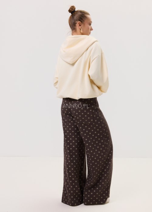 Brązowe damskie spodnie Retro Dots z szeroką nogawką, ozdobione beżowymi groszkami. Model z wysokim stanem i paskiem w talii, w stylizacji z kremowym topem oraz brązowymi botkami.