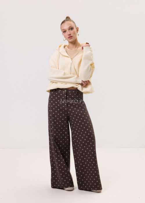 Brązowe damskie spodnie Retro Dots z szeroką nogawką, ozdobione beżowymi groszkami. Model z wysokim stanem i paskiem w talii, w stylizacji z kremowym topem oraz brązowymi botkami.