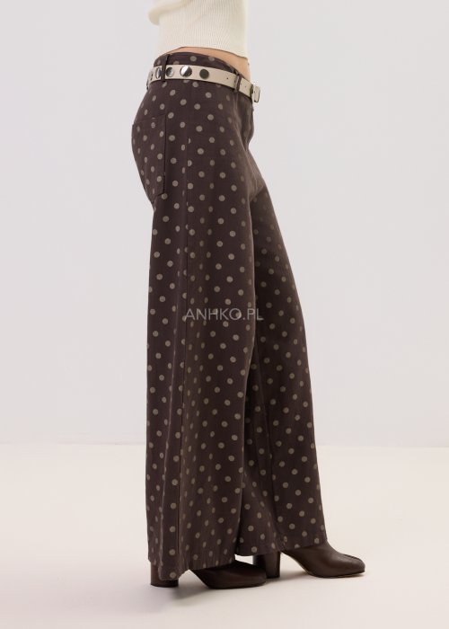 Brązowe damskie spodnie Retro Dots z szeroką nogawką, ozdobione beżowymi groszkami. Model z wysokim stanem i paskiem w talii, w stylizacji z kremowym topem oraz brązowymi botkami.