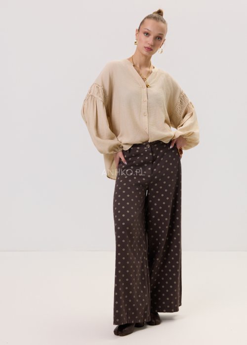 Brązowe damskie spodnie Retro Dots z szeroką nogawką, ozdobione beżowymi groszkami. Model z wysokim stanem i paskiem w talii, w stylizacji z kremowym topem oraz brązowymi botkami.