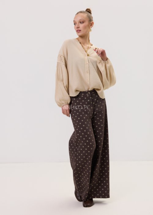 Brązowe damskie spodnie Retro Dots z szeroką nogawką, ozdobione beżowymi groszkami. Model z wysokim stanem i paskiem w talii, w stylizacji z kremowym topem oraz brązowymi botkami.