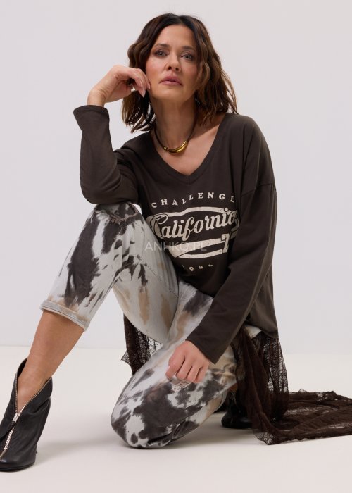 Brązowa bluzka oversize z długim rękawem i dekoltem V, z nadrukiem „California 79”, zestawiona z jeansami tie-dye i koronkową narzutką w stylizacji casual