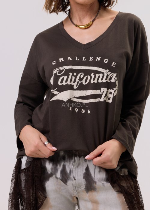 Brązowa bluzka oversize z długim rękawem i dekoltem V, z nadrukiem „California 79”, zestawiona z jeansami tie-dye i koronkową narzutką w stylizacji casual