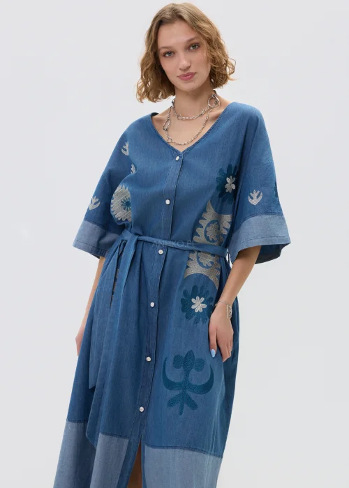 Jeansowa sukienka maxi w odcieniu niebieskiego denimu z haftowanymi wzorami, wiązana w talii paskiem, zapinana na guziki, o luźnym fasonie z szerokimi rękawami, stylizowana z kowbojkami.