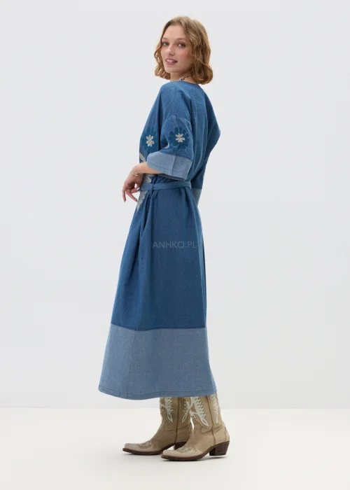 Jeansowa sukienka maxi w odcieniu niebieskiego denimu z haftowanymi wzorami, wiązana w talii paskiem, zapinana na guziki, o luźnym fasonie z szerokimi rękawami, stylizowana z kowbojkami.