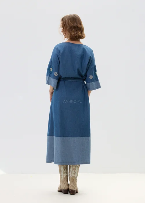 Jeansowa sukienka maxi w odcieniu niebieskiego denimu z haftowanymi wzorami, wiązana w talii paskiem, zapinana na guziki, o luźnym fasonie z szerokimi rękawami, stylizowana z kowbojkami.