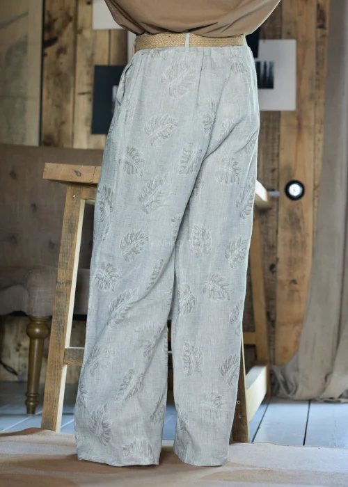Lniane spodnie w szarym kolorze z subtelnym roślinnym wzorem, o szerokiej nogawce i luźnym kroju, stylizowane z paskiem w naturalnym odcieniu w minimalistycznym, eleganckim looku.