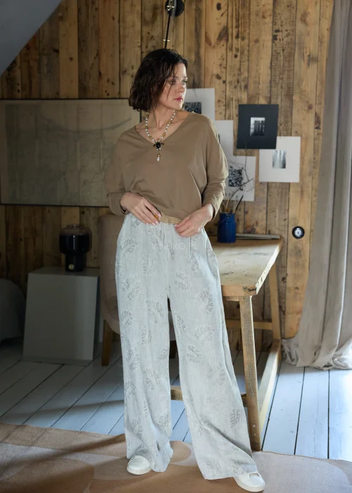 Lniane spodnie w szarym kolorze z subtelnym roślinnym wzorem, o szerokiej nogawce i luźnym kroju, stylizowane z paskiem w naturalnym odcieniu w minimalistycznym, eleganckim looku.