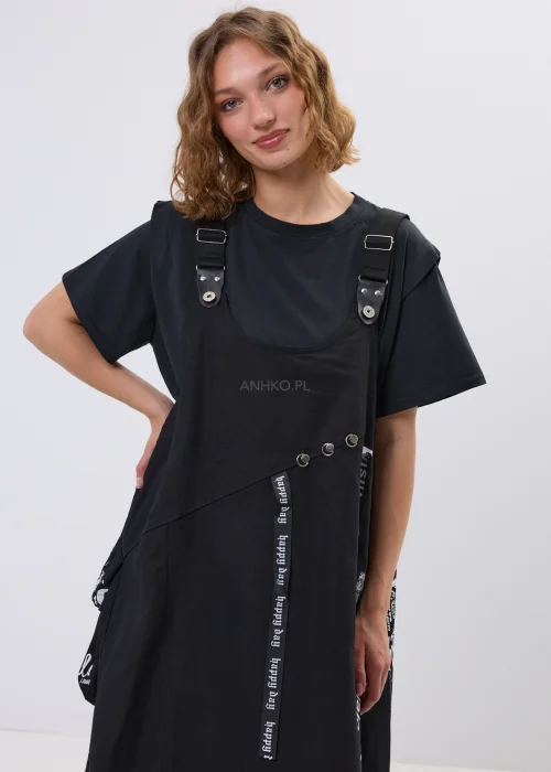 Czarna sukienka ogrodniczka oversize z ozdobnymi taśmami i metalowymi detalami w nowoczesnej miejskiej stylizacji.