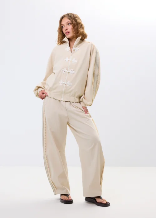Beżowa bluza oversize na zamek z ozdobnymi wiązaniami i wysokim kołnierzem. Luźny fason nadaje stylizacji nowoczesny i oryginalny charakter.