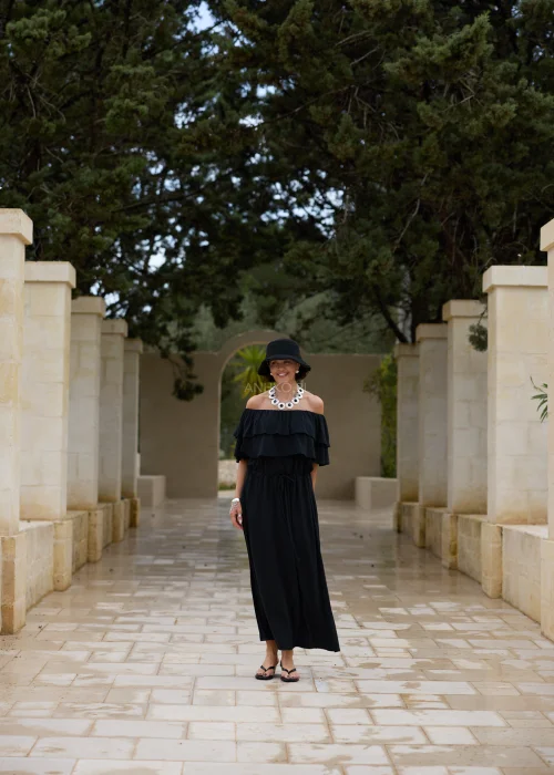 Czarna sukienka maxi hiszpanka z efektownymi falbanami przy dekolcie i regulacją w talii. Elegancka stylizacja z odkrytymi ramionami, kapeluszem i wyrazistą biżuterią w minimalistycznym wydaniu.