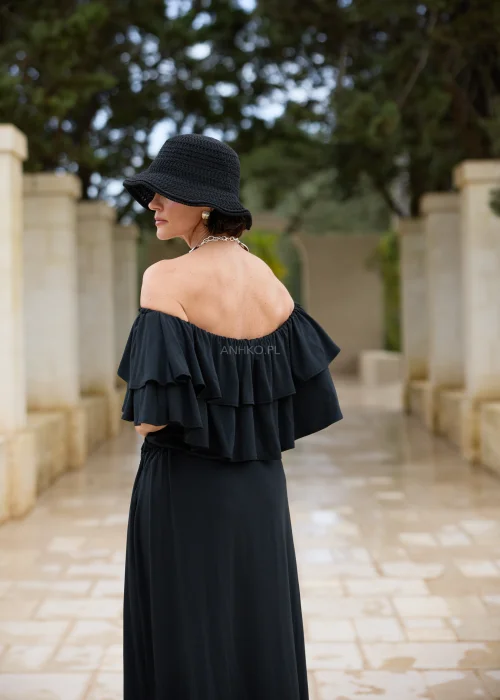 Czarna sukienka maxi hiszpanka z efektownymi falbanami przy dekolcie i regulacją w talii. Elegancka stylizacja z odkrytymi ramionami, kapeluszem i wyrazistą biżuterią w minimalistycznym wydaniu.