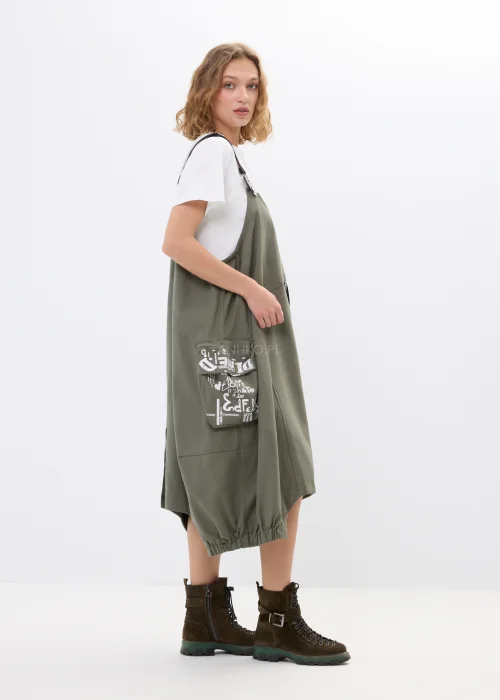Khaki sukienka ogrodniczka z nadrukami, dużymi kieszeniami i regulowanymi szelkami. Luźny model w miejskim stylu z nowoczesnymi detalami i asymetrycznym fasonem.