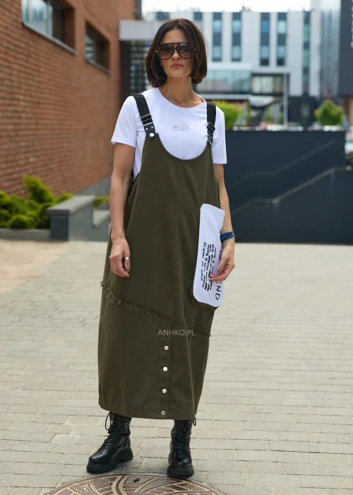 Khaki sukienka ogrodniczka midi z regulowanymi szelkami i kontrastową kieszenią z nadrukiem. Casualowy model o luźnym fasonie, zestawiony z białym T-shirtem i czarnymi botkami.