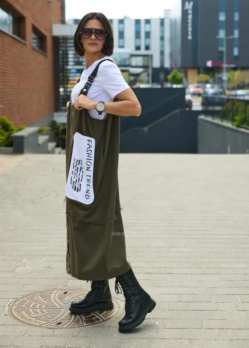 Khaki sukienka ogrodniczka midi z regulowanymi szelkami i kontrastową kieszenią z nadrukiem. Casualowy model o luźnym fasonie, zestawiony z białym T-shirtem i czarnymi botkami.