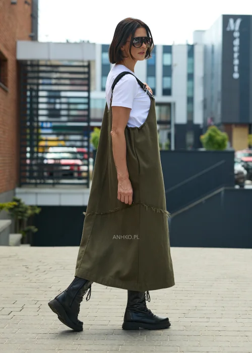 Khaki sukienka ogrodniczka midi z regulowanymi szelkami i kontrastową kieszenią z nadrukiem. Casualowy model o luźnym fasonie, zestawiony z białym T-shirtem i czarnymi botkami.