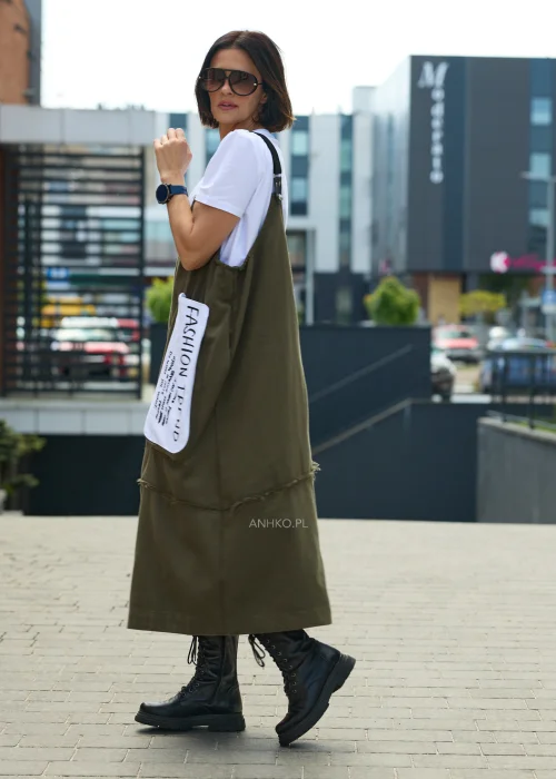 Khaki sukienka ogrodniczka midi z regulowanymi szelkami i kontrastową kieszenią z nadrukiem. Casualowy model o luźnym fasonie, zestawiony z białym T-shirtem i czarnymi botkami.