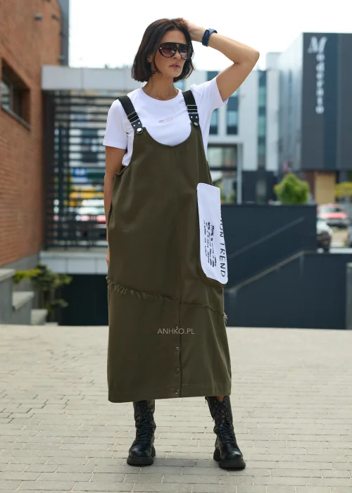 Khaki sukienka ogrodniczka midi z regulowanymi szelkami i kontrastową kieszenią z nadrukiem. Casualowy model o luźnym fasonie, zestawiony z białym T-shirtem i czarnymi botkami.