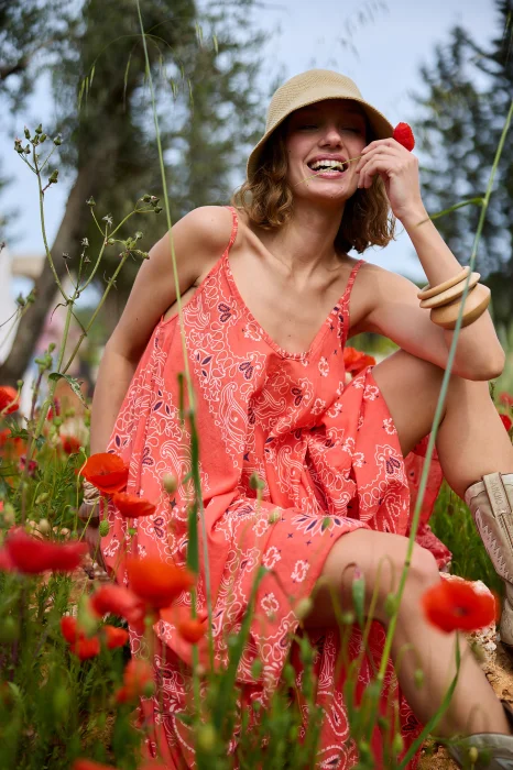 Koralowa sukienka maxi na cienkich ramiączkach z ozdobnym wzorem paisley. Zwiewny model o rozkloszowanym kroju prezentowany w stylizacji boho z kapeluszem i kowbojkami.