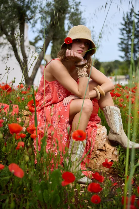 Koralowa sukienka maxi na cienkich ramiączkach z ozdobnym wzorem paisley. Zwiewny model o rozkloszowanym kroju prezentowany w stylizacji boho z kapeluszem i kowbojkami.