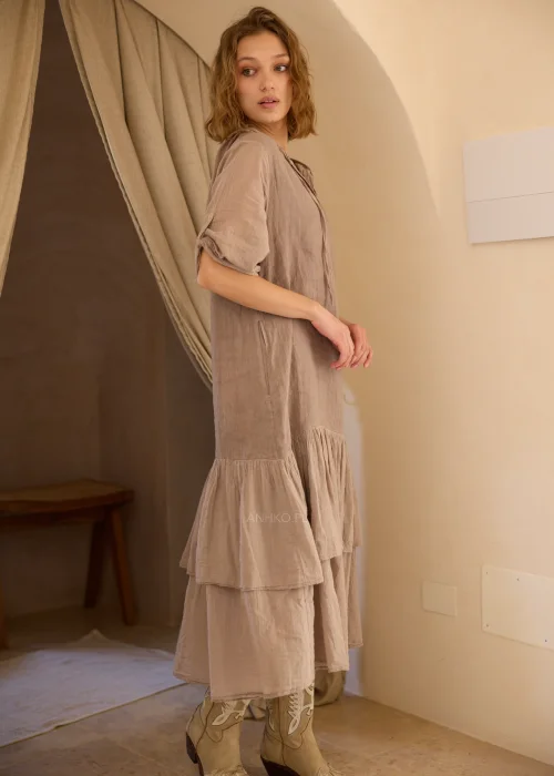Lniana sukienka maxi z kapturem w odcieniu taupe, z dekoracyjną falbaną u dołu i podwijanymi rękawami. Swobodny fason w stylu boho doskonale sprawdzi się w naturalnych, codziennych i eleganckich stylizacjach.