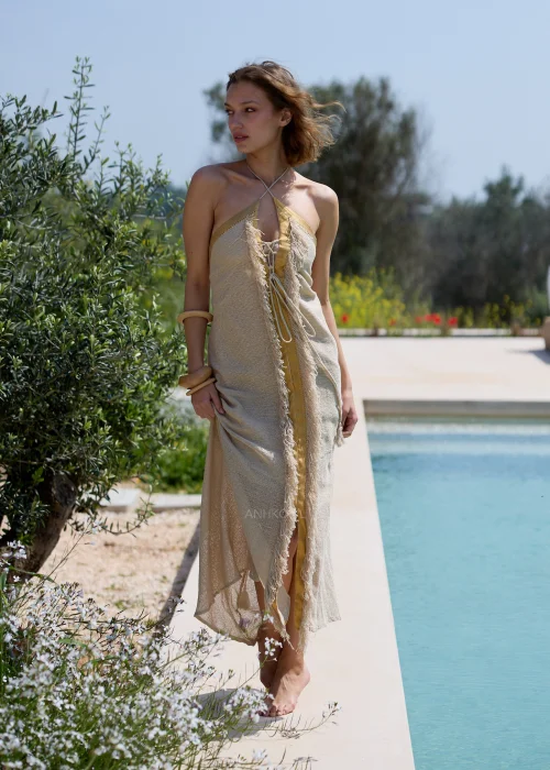 Beżowa sukienka maxi w stylu boho z wiązaniem przy dekolcie, odkrytymi ramionami i ozdobnymi frędzlami. Zwiewny model o naturalnym charakterze, idealny na wakacyjne stylizacje i wypoczynek nad wodą.