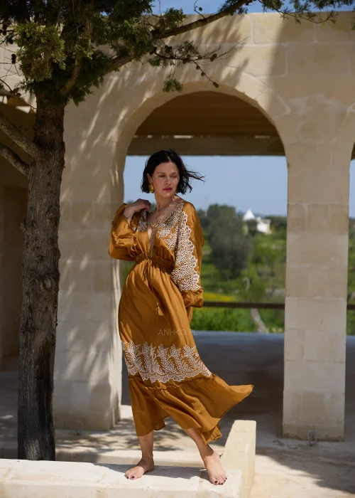 Karmelowa sukienka boho midi z ozdobną koronką i haftowanymi wstawkami przy dekolcie, rękawach oraz dole. Kobiecy fason z wiązaniem w talii i falbaną tworzy elegancką stylizację w klimacie śródziemnomorskim.