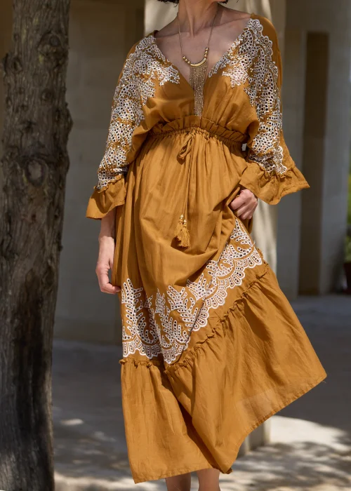 Karmelowa sukienka boho midi z ozdobną koronką i haftowanymi wstawkami przy dekolcie, rękawach oraz dole. Kobiecy fason z wiązaniem w talii i falbaną tworzy elegancką stylizację w klimacie śródziemnomorskim.