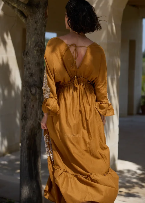 Karmelowa sukienka boho midi z ozdobną koronką i haftowanymi wstawkami przy dekolcie, rękawach oraz dole. Kobiecy fason z wiązaniem w talii i falbaną tworzy elegancką stylizację w klimacie śródziemnomorskim.