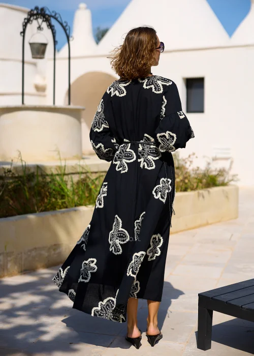 Czarna sukienka maxi z efektownymi białymi haftami w kształcie kwiatów, bufiastymi rękawami i ozdobnym paskiem w talii. Zwiewny model w stylu boho, idealny na eleganckie i codzienne stylizacje z kobiecym charakterem.